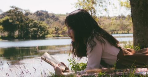 A teenage girl with black hair reads a book while laying on the grass by a stream